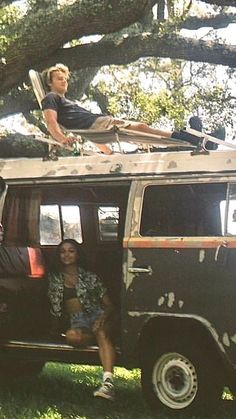 two people sitting on the roof of an old vw van under a large tree