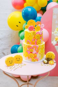 there is a yellow cake with smiley faces on it and balloons in the back ground