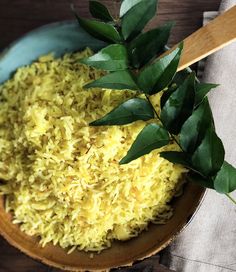 a wooden bowl filled with yellow rice next to a green leafy branch on top of a table