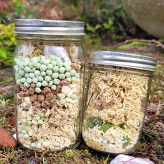 two mason jars filled with noodles and other things sitting on the ground next to rocks