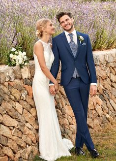 a bride and groom standing next to a stone wall with lavenders in the background