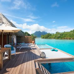 an outdoor deck with chairs and umbrellas next to the water in front of a thatched hut