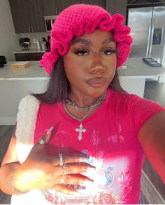 a woman wearing a pink hat and holding her hand up to the camera while standing in a kitchen