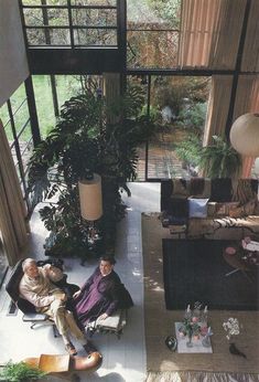 two men sitting on the floor in a living room with large windows and potted plants