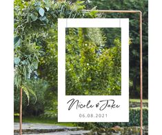 a wedding sign hanging from a metal frame with greenery in the back ground and trees behind it