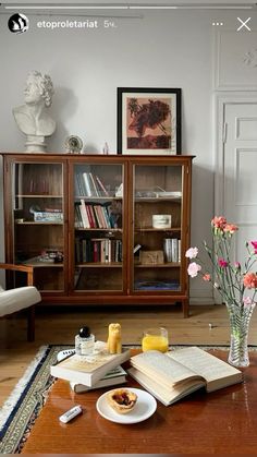 an open book on a table with flowers in a vase and bookshelf behind it