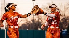 two female softball players in orange uniforms are holding their mitts up to each other