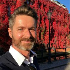 a man standing in front of a building with red leaves on the trees behind him