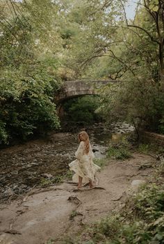 a woman in a white dress walking down a path near a river with trees and a bridge