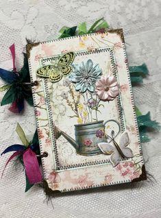 a small card with flowers and a watering can on it's side, sitting on a lace tablecloth