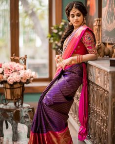 a woman in a purple and pink saree leaning on a wall with flowers behind her