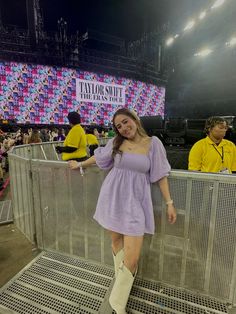 a woman standing in front of a metal fence at a music festival wearing boots and a purple dress