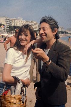 a man standing next to a woman holding a cell phone on top of a beach