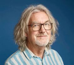 an older man wearing glasses and a striped shirt looks at the camera while standing in front of a blue background