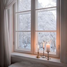 two candles sit on a window sill in front of the snow - covered trees