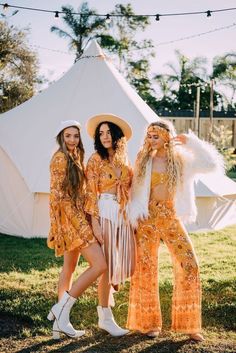 three women in matching outfits standing next to each other near a white tent with string lights