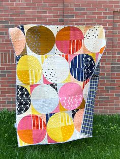 a patchwork quilt hanging on the side of a brick building with grass in front of it