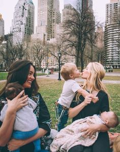 two women and a baby are sitting on the grass in front of tall buildings with skyscrapers behind them