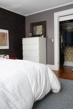 a white bed sitting in a bedroom next to a dresser and closet with clothes hanging on it