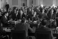 a group of men sitting around a table in front of a microphone and people standing behind them