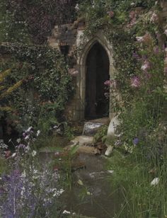 an old stone building surrounded by flowers and greenery with a stream running through it