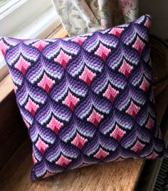 a purple and pink knitted pillow sitting on top of a wooden table next to a window