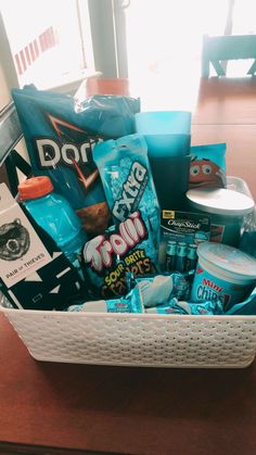 a white basket filled with snacks and drinks on top of a wooden table next to a window