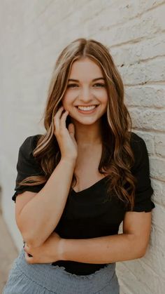 a woman leaning against a white brick wall talking on her cell phone and smiling at the camera