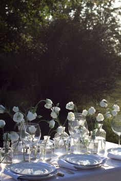 white flowers are in glass vases and plates on a table with trees in the background