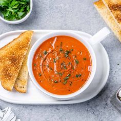 a white plate topped with a bowl of tomato soup next to a slice of bread