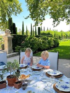 two young children sitting at a table with plates and bowls on it in front of some trees