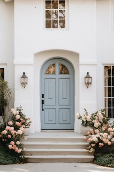 a blue front door on a white house with flowers in the foreground and two large windows above it