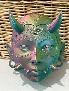 a green and purple mask with horns on it's head sitting next to a wicker basket