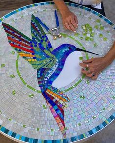 a bird made out of glass sitting on top of a mosaic table with a person working on it