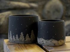 two black cups sitting on top of a wooden table next to a stone wall with snow covered mountains in the background