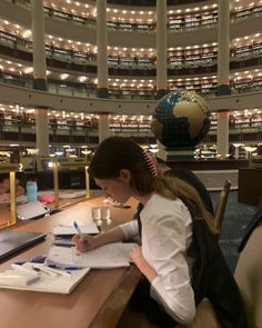 a woman sitting at a table writing in a notebook with a globe on the wall behind her