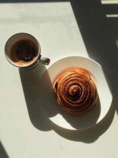 a white plate topped with a cinnamon roll next to a cup of coffee on top of a table