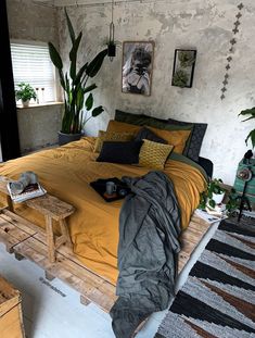 a bed with yellow sheets and pillows in a room next to a plant filled wall