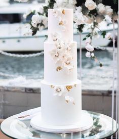 a three tiered white wedding cake with flowers on the top and bottom, sitting on a glass table