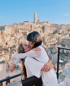 two women hugging each other on top of a hill with buildings in the background and blue sky