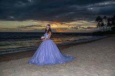 a woman in a purple dress standing on the beach at sunset with palm trees behind her