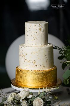 a white and gold wedding cake sitting on top of a table next to greenery