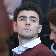 a man in a red sweater and white collared shirt looks to his left as he waits for the judge