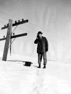 a man standing in the snow talking on a cell phone next to a telephone pole