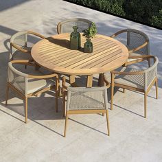 an outdoor table with six chairs and two vases on the top, in front of some bushes