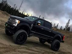 a black truck driving down a dirt road under a cloudy sky with trees in the background