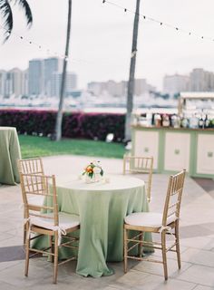 the table is set with two chairs and a green tablecloth on it in front of some palm trees