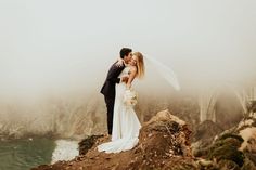 a bride and groom kissing on top of a cliff overlooking the ocean in foggy weather