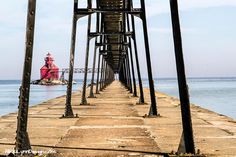 a long pier with a red lighthouse in the distance and water behind it, on a sunny day