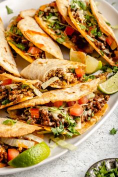 several tacos on a white plate with limes and cilantro in the background